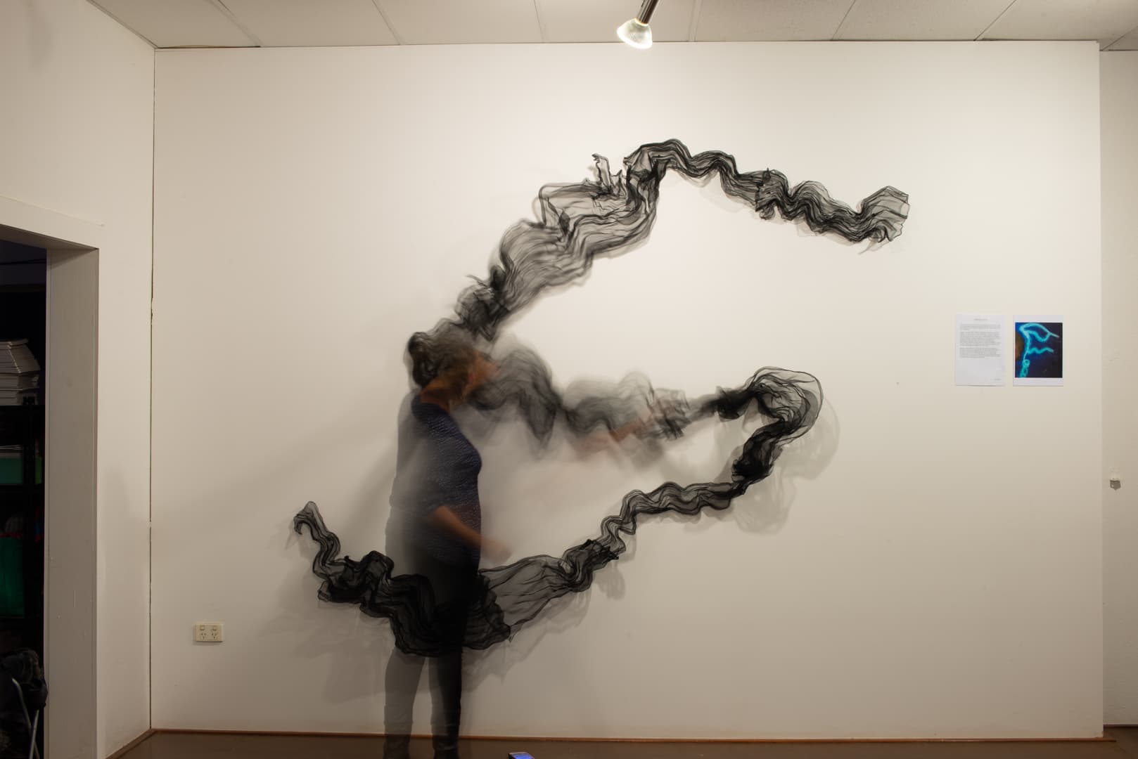 Blurred figure standing in front of a large black mesh wall installation by J9 Stanton.